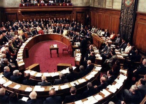 Inside a council meeting, showing three rings of seats and desks filled with mostly male Councillors.