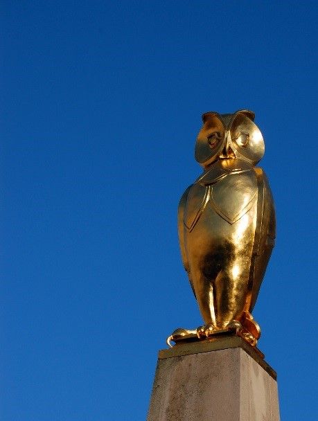 A beautifully crafted golden owl, standing tall with its wings tucked in against a clear blue sky.