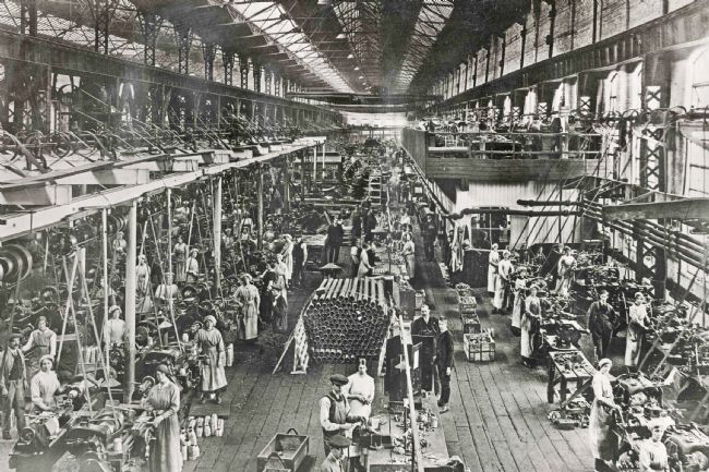 Wide view of the factory with many workers, rows of machines and piles of shell cases.