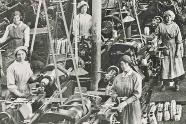 Women in factory uniforms standing by heavy machinery with empty shell cases on the floor.