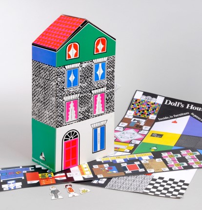 A photo of the cardboard press-out doll's house designed by Maureen Roffey. This image shows a brightly coloured cardboard house which stands at around nine inches tall. The design includes a red and pink patterned roof, some brick pattern on the external walls, seven windows and a front door. Lying beneath the stood-up house are some of the pages which have not yet been pressed out, including furniture to be assembled, and some of the flooring and wall designs.