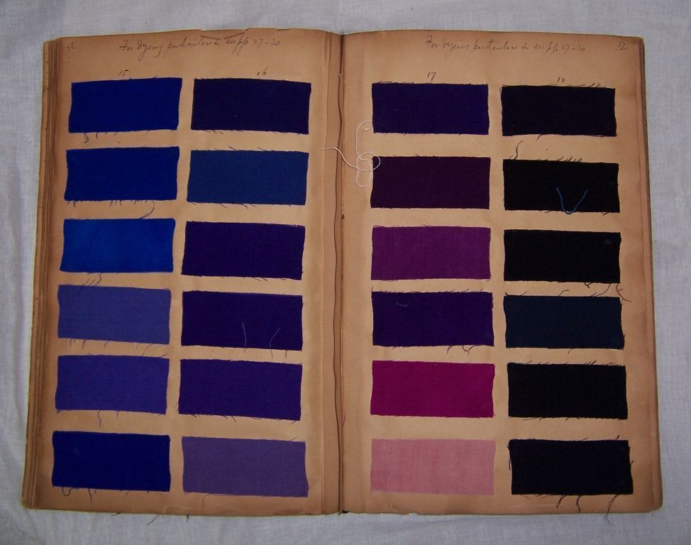 Open pages of an old dye book displaying fabric swatches. The left page features six pairs of blue and purple fabric samples, neatly arranged in two columns. The right page shows six pairs of purple, pink, and black fabric samples. The pages are yellowed with age, and handwritten notes are visible at the top of both pages.