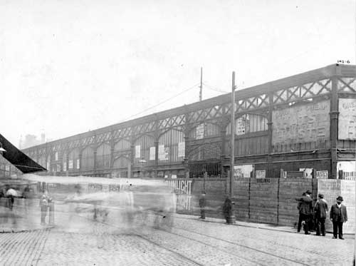 Black and white photograph showing one side of Kirkgate market.  There is a small group of men wearing suits and hats standing on the corner