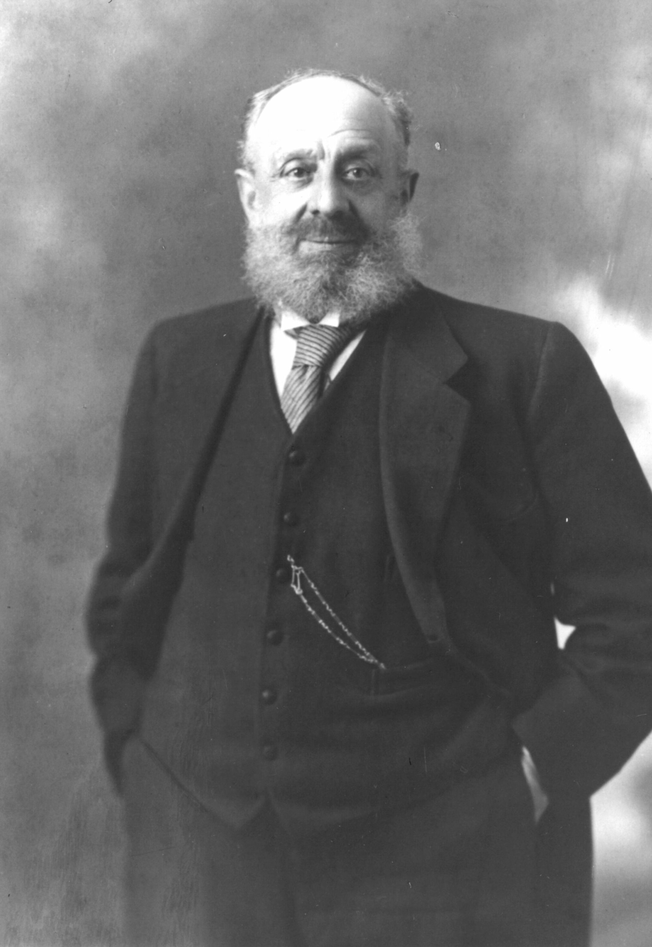 Black and white portrait photo of elderly bearded man wearing a suit and tie, standing with hands in pockets