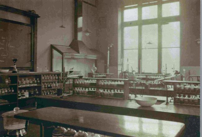 Photo of an 1890s classroom at Huddersfield Mechanical Institute showing desks and rows of glass bottles on shelves.