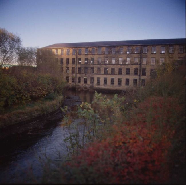 Photograph of Armley Mills showing a long building with multiple windows set next to a river.