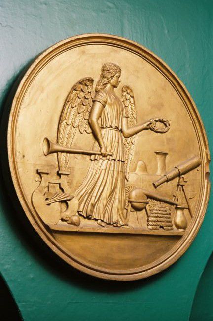 Dark gold coloured plaque with an angel holding a trumpet, surrounded by items from industry, such as a telescope and a bee-hive