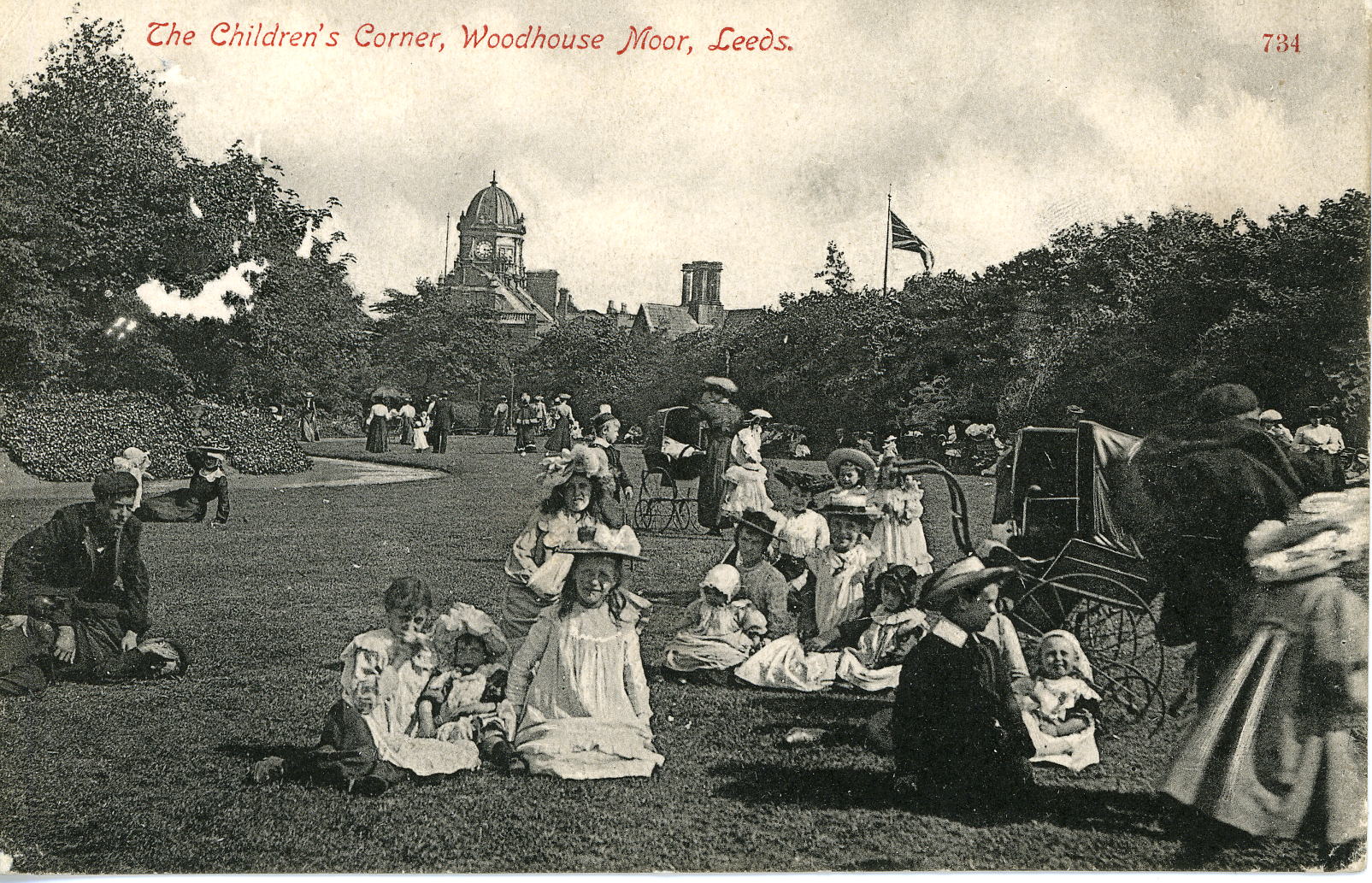 Black and white photograph of groups of families sitting on the grass at Woodhouse Moor. In the foreground there are three main family groups, each with between three and six children. The girls are dressed in long white aprons and straw hats, the boys in suits and boaters. In the background a path winds to the left and there are trees and buildings.