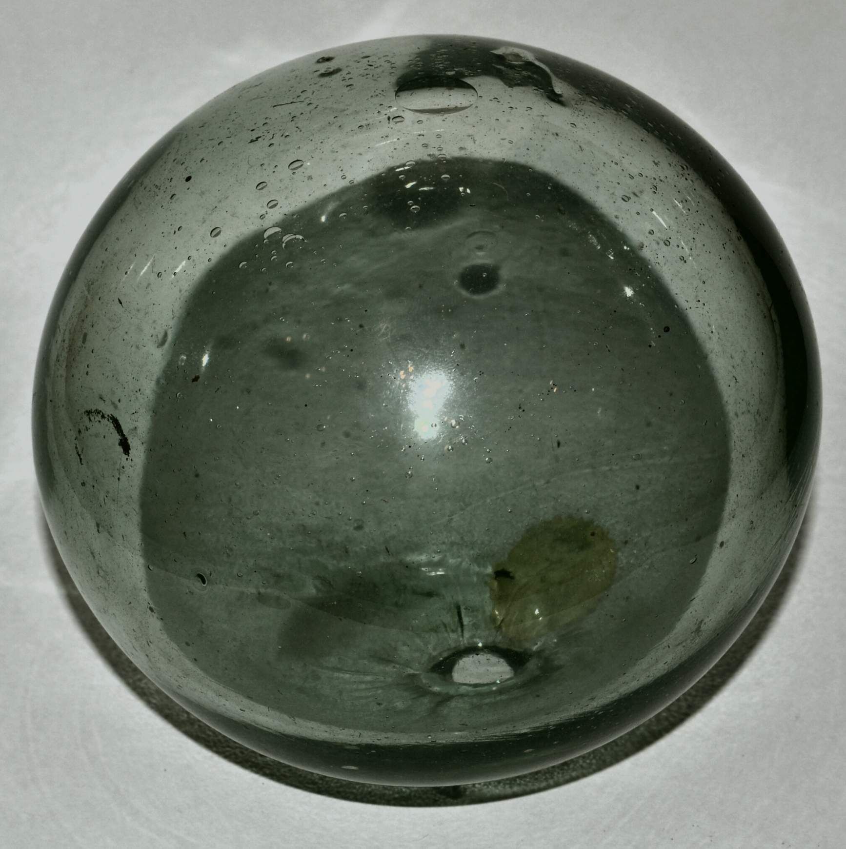 A greyish translucent glass ball with air bubbles and surface imperfections sits on a white surface, reflecting light slightly.