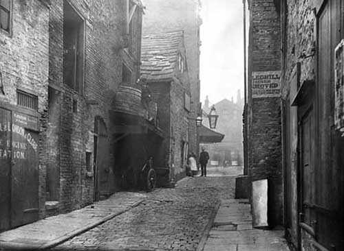 Black and white photograph showing buidings on either side of a cobbled street.   There are no drains.