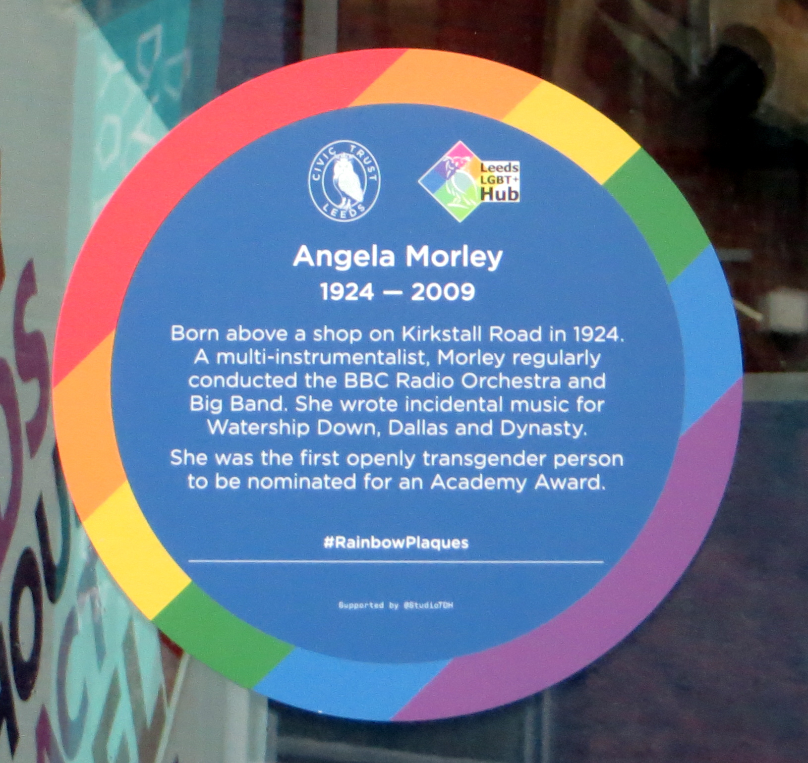 Colour photograph showing the sticker of a rainbow edged plaque dedicated to Angela Morley