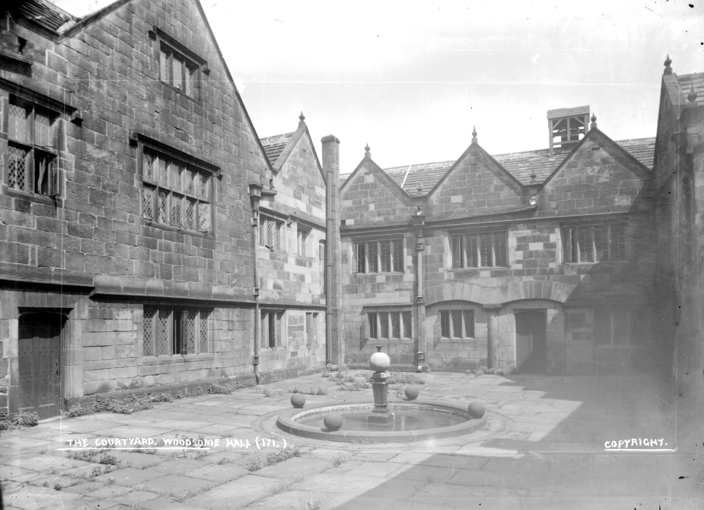 A black and white photo of a historic stone courtyard at Woodsome Hall with medieval architecture. Features include mullioned windows and a central fountain.