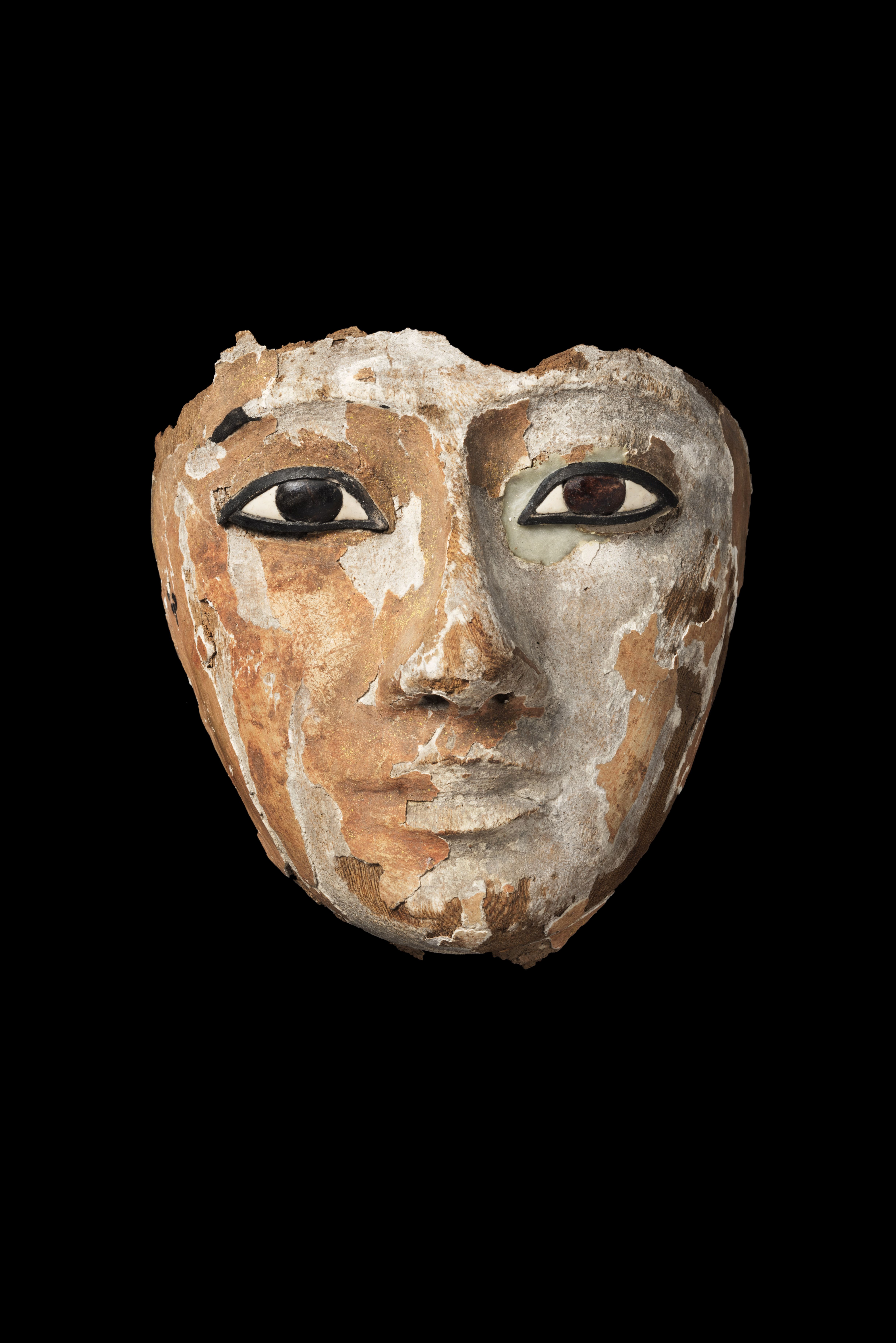 A worn ancient Egyptian coffin face mask, featuring large, almond-shaped eyes with dark, outlined rims. The face has a serene expression, though much of the surface has flaked away, revealing patches of the original material beneath. The remaining paint is faded, with a mix of beige, brown, and white tones. The mask's weathered appearance reflects its age and long history.
