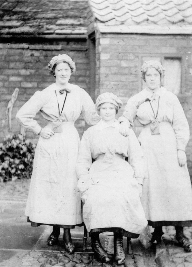 Black and white photograph of three young female factory workers wearing overalls.  They are in skirts rather than trousers.