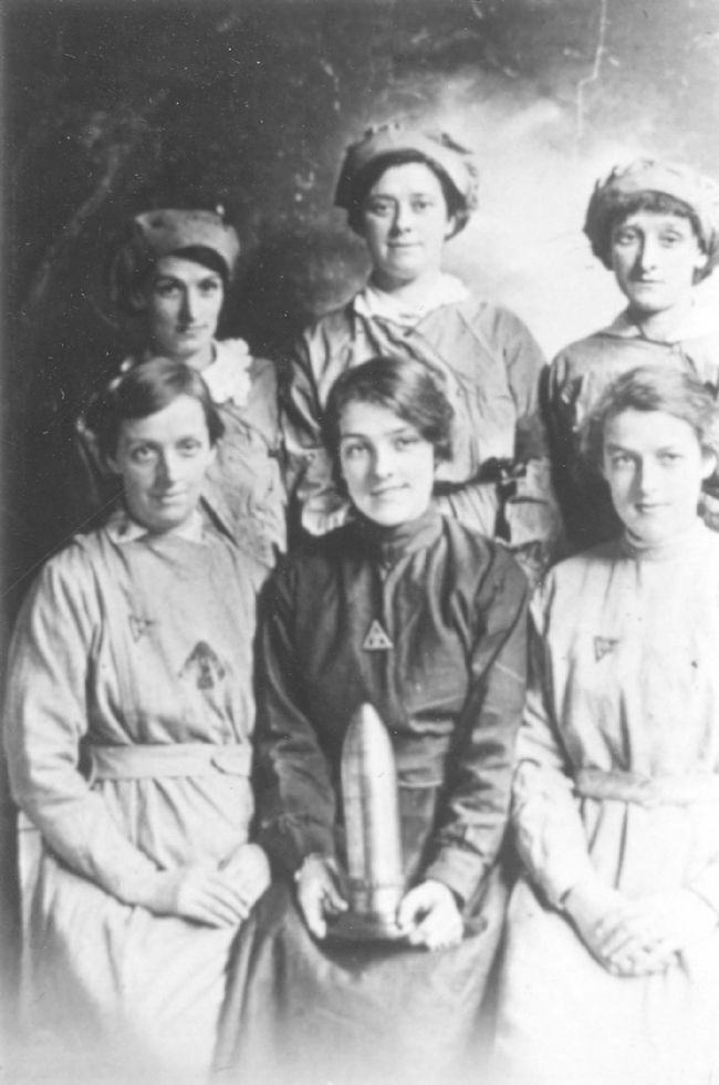 Black and white photograph of six female factory workers. The girl in the front, centre, is holding a shell case.