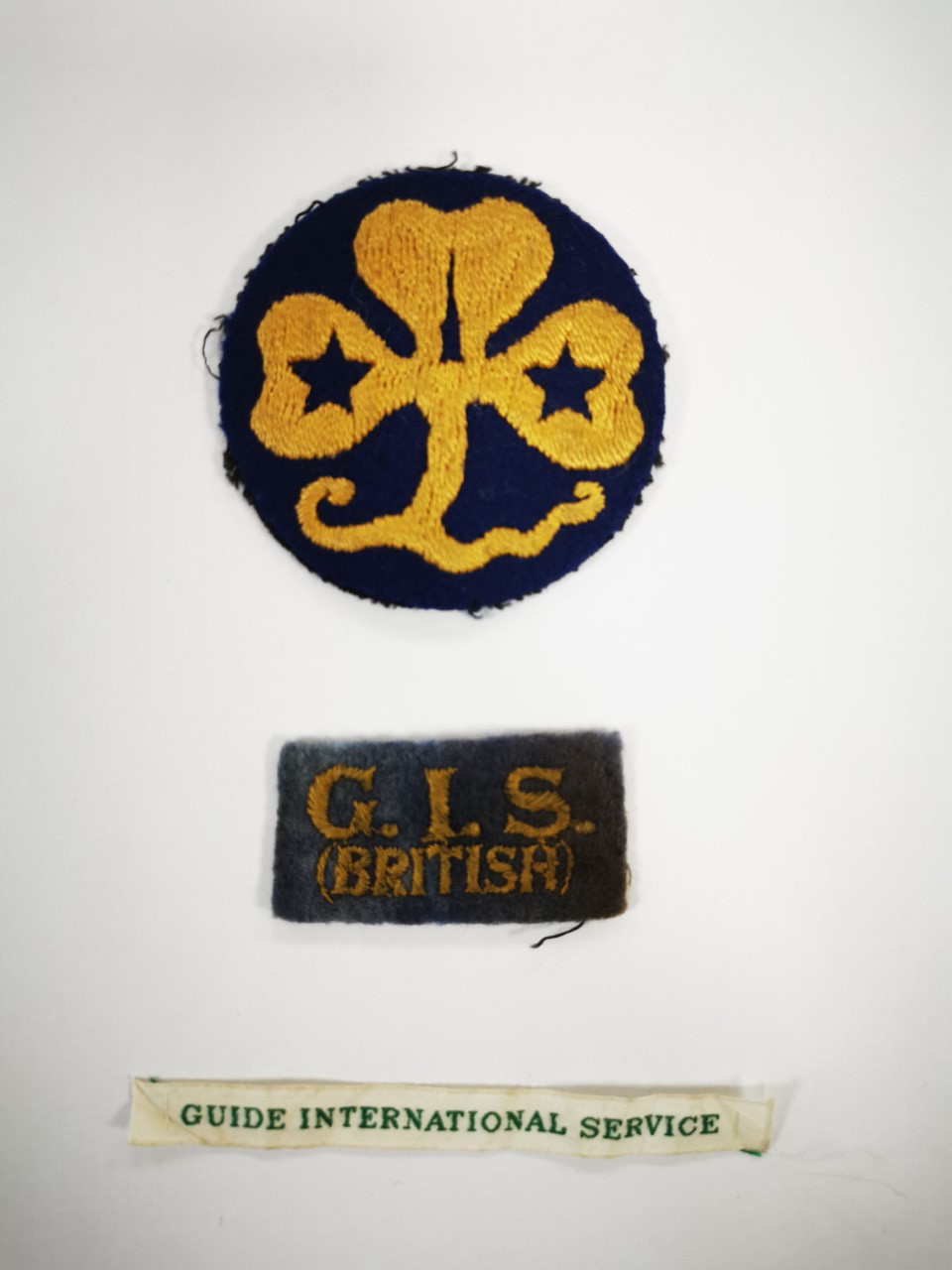 Three badges worn by Girl Guides on International Service - a circular trefoil one, one with GIS (British) on and one that says 'Guide International Service'