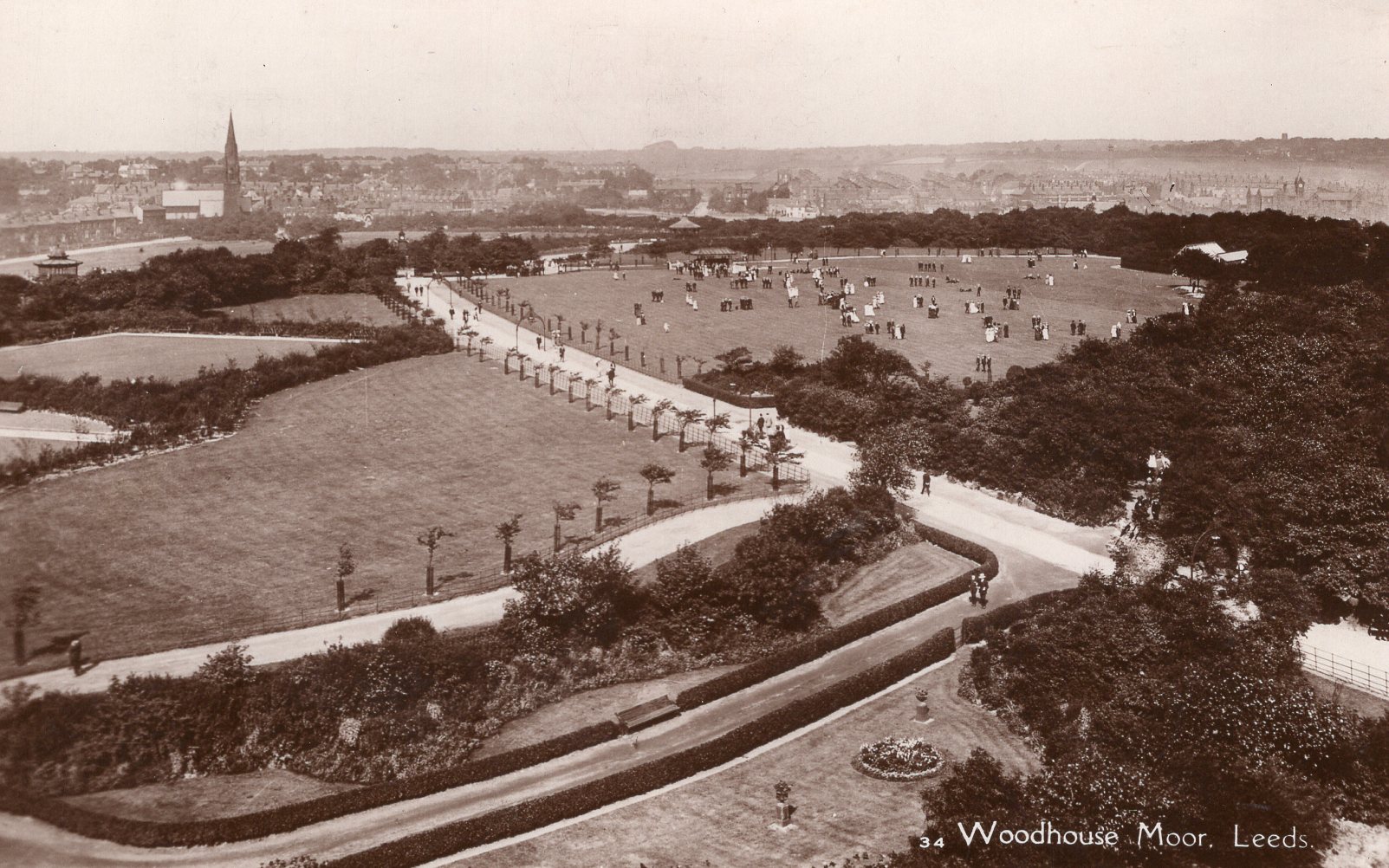 Black and white photograph showing a view of Woodhouse Moor park from above. There are aeras of grass, split with paths and trees criss-crossing the space. Some people can be seen on the paths. There are houses and buildings in the background