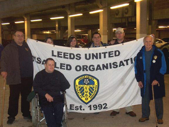 Colour photograph showing a group of people holding a flag which has Leeds United Disabled Organisation printed on it.