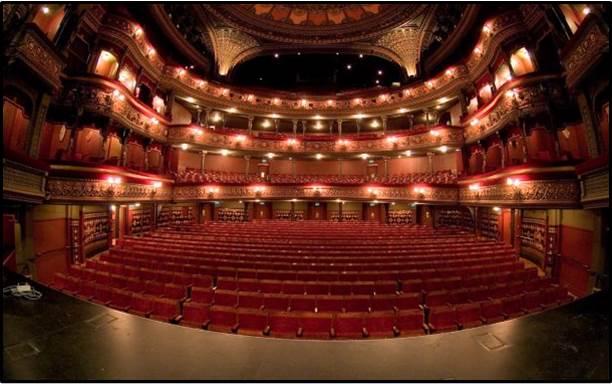 The theatre is empty, with lights on the three tiers of balconies.  The seats are upholstered in red.