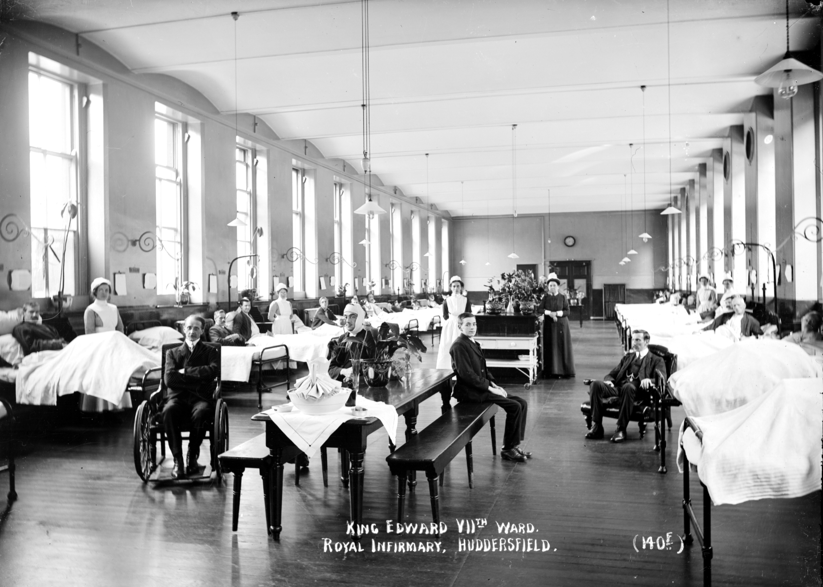 A black and white photograph of a historic hospital ward with rows of beds, patients seated, and nurses attending. The room has large windows and a high ceiling.