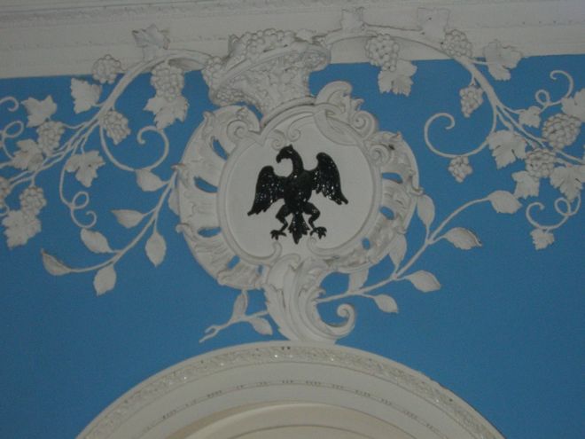 Close-up photograph showing a blue wall with white plasterwork of a grape vine and an eagle.  The eagle is painted black.