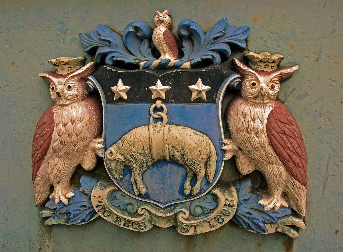 The Leeds coat of arms has a central blue shield with three white stars across the top.  Under these is a sheep.  On either side of the shield is a large owl, with a smaller one on top.  The latin motto is written on a red banner underneath.