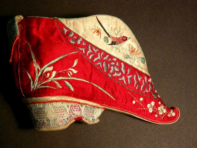 The shoe is made from red and white satin with embroidered images of a bird and flowers.  It has a  small heel and ends in a sharp point at the toe end.  It is about 3 inches long.