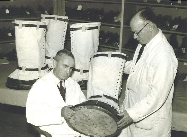 Two men holding one of the elephant boots.  The boot is about as long as one of their arms and much, much wider.
