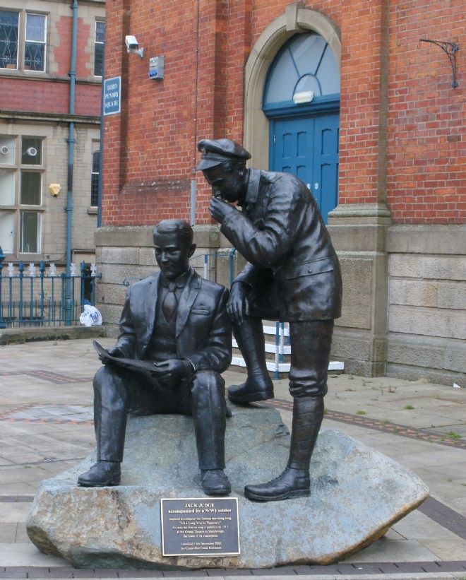 Bronze statue of a man whispering in a seated man's ear, while he writes something. Plaque labelled 'Jack Judge below.