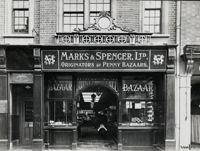 Black and white photograph showing the front of an M&S store. It say's 'Bazaar' in the windows each side of the big double door.