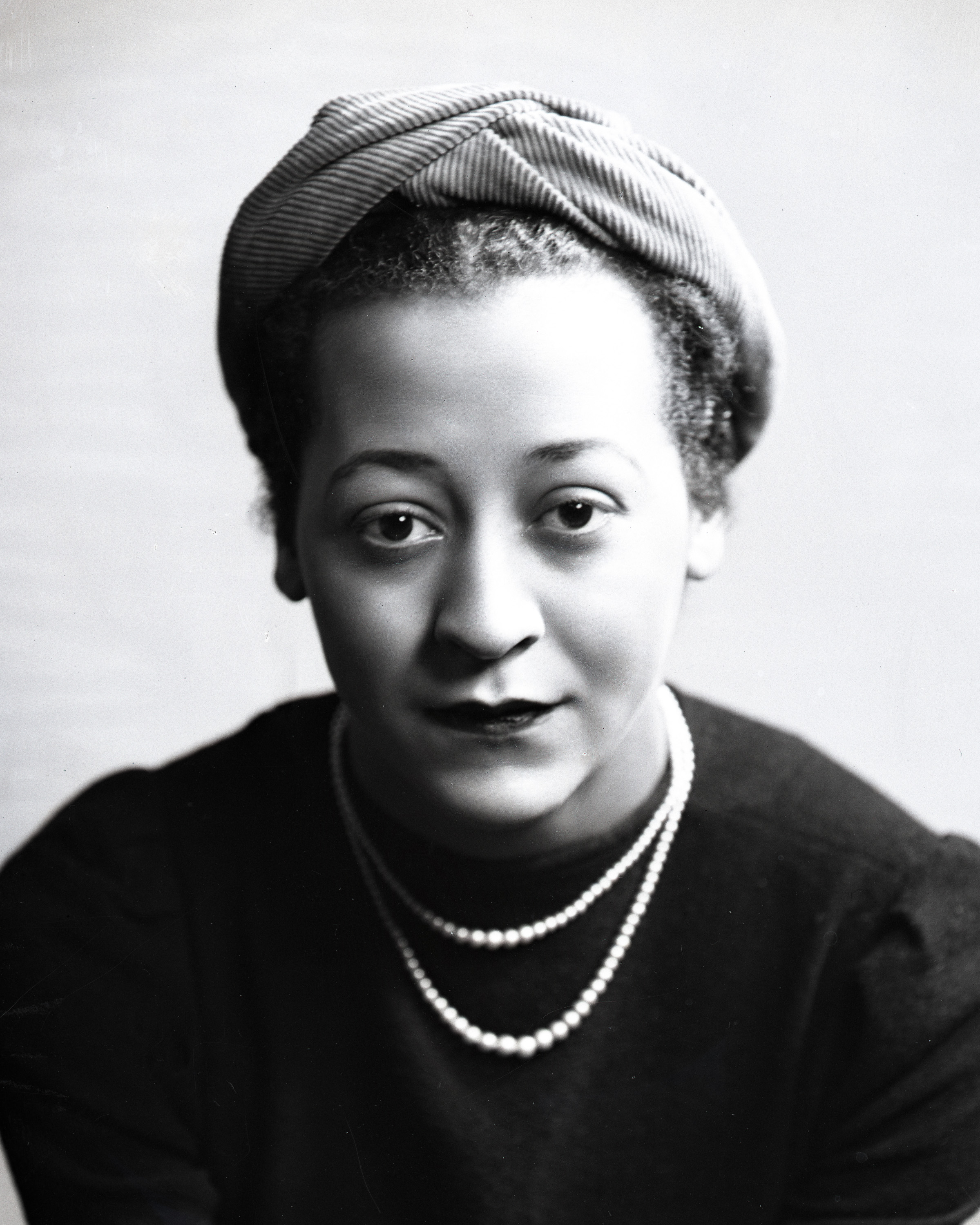 Black and white portrait photo of a Black woman wearing a scarf on her head, a dark coloured top and a white necklace