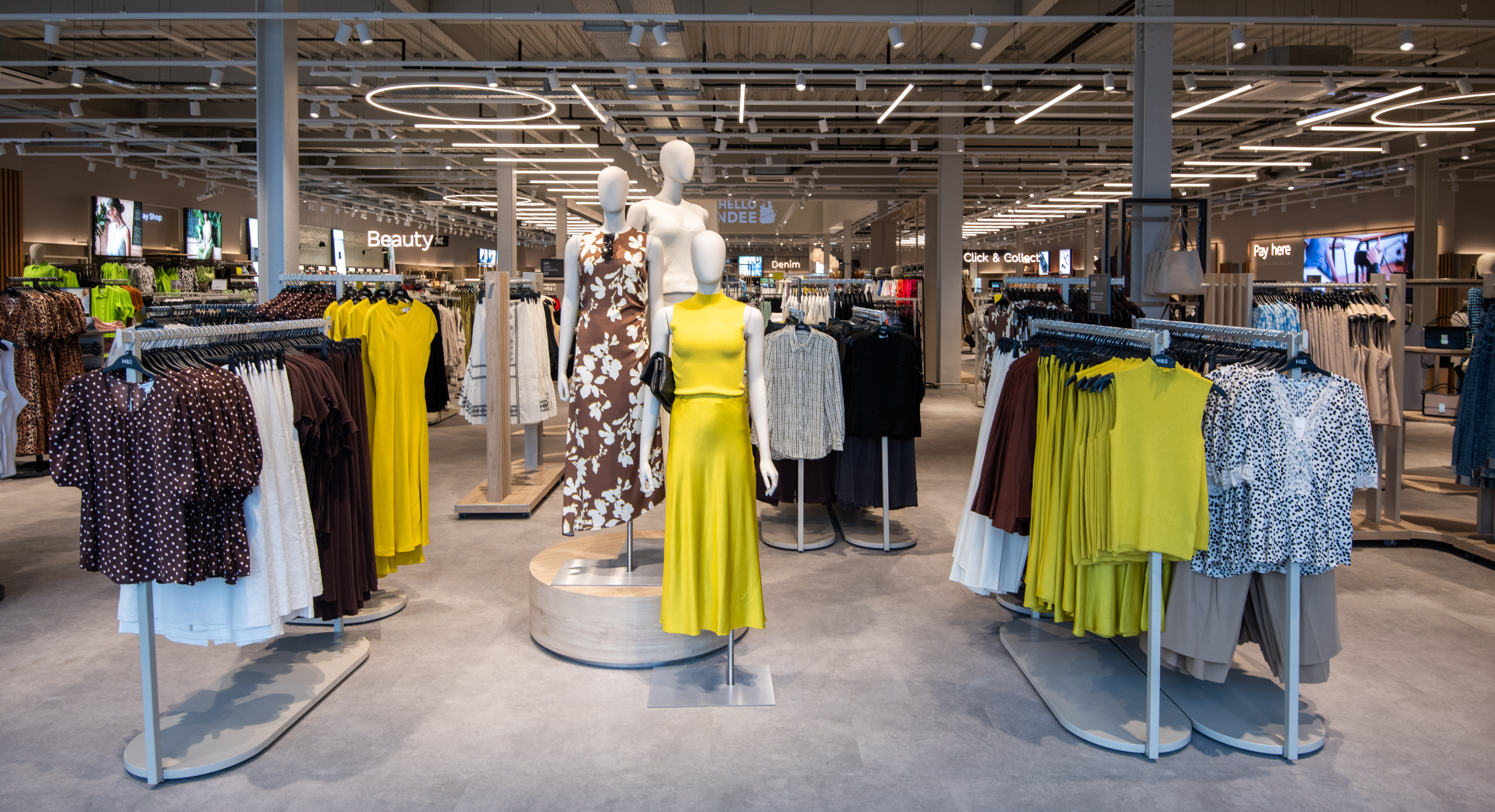 Marks & Spencer store interior with modern lighting. Three mannequins display vibrant dresses in yellow and floral patterns, surrounded by colourful clothing racks.