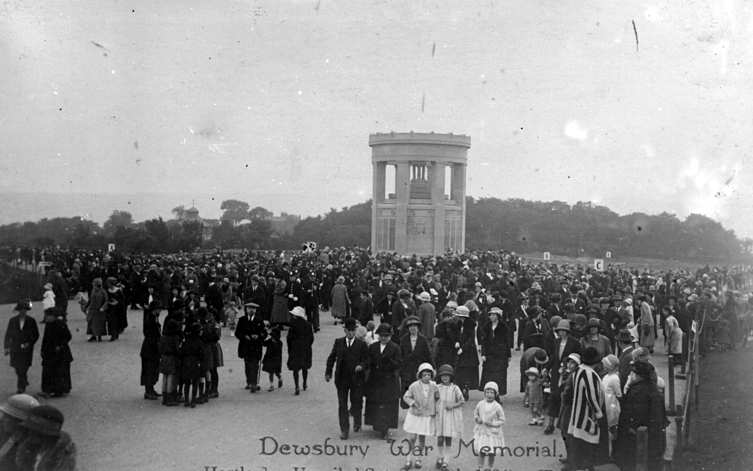 A black and white photograph of a crowd gathered around the Dewsbury War Memorial, an imposing stone structure with pillars, set in a spacious park.