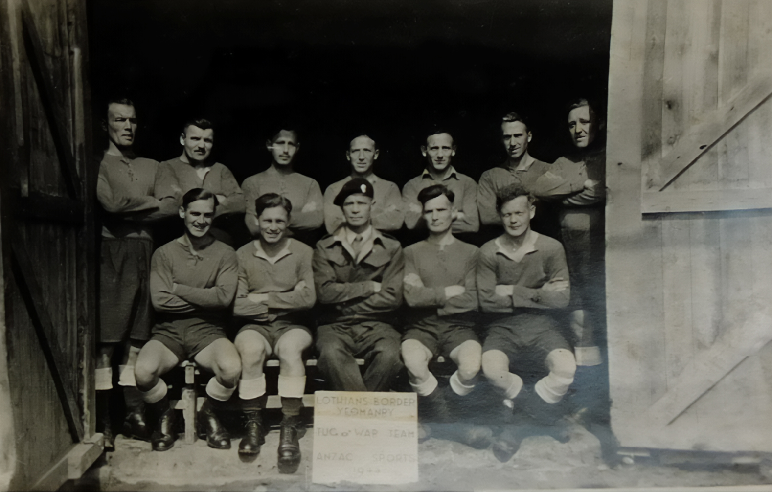 Black and white photo of a POW tug of war team posed in rows inside a wooden barn or shed.