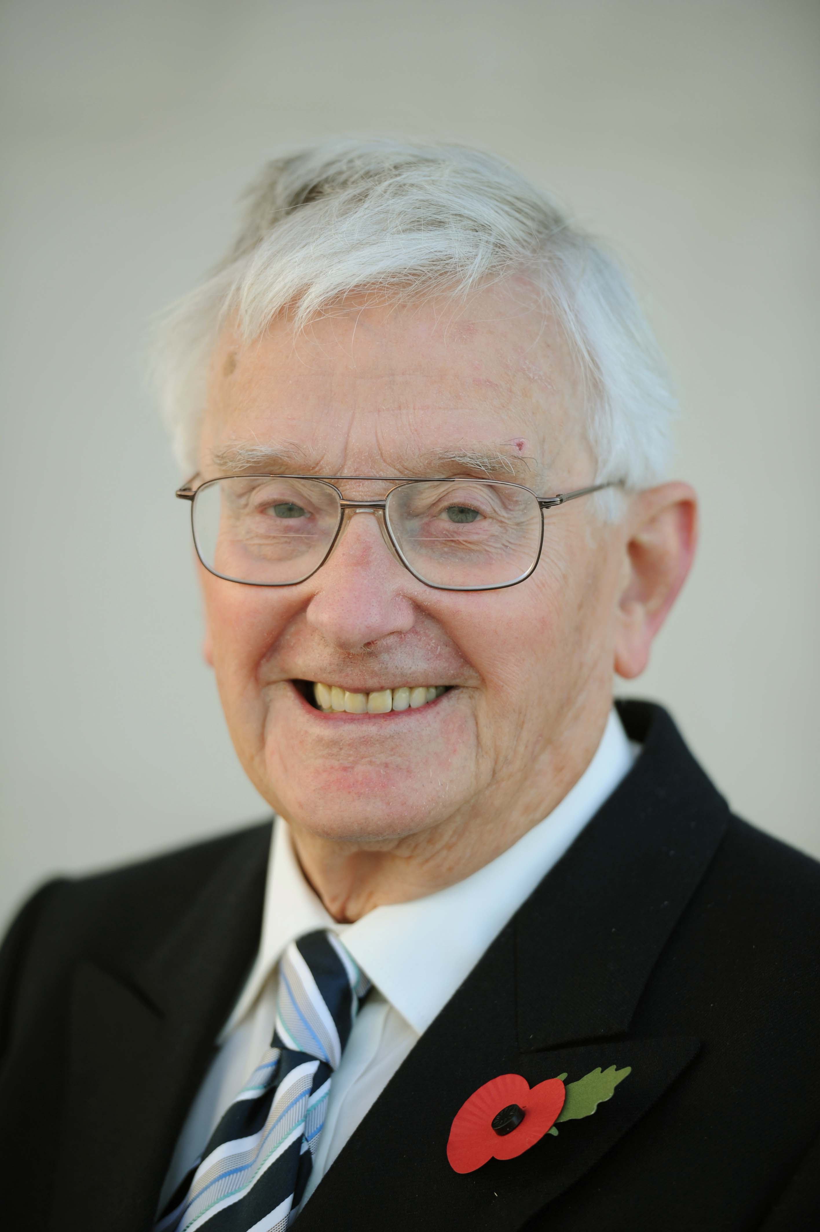 Colour photograph of an older man wearing a suit jacket with a poppy. He has white hair and is wearing glasses.
