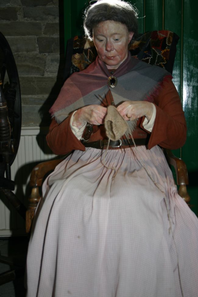 Model of woman sitting in a rocking chair knitting. She is wearing a short scarf covering her shoulders which is pinned at the front with a brooch.