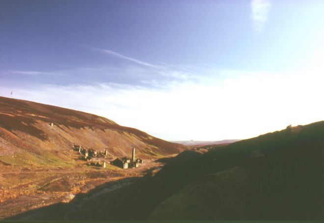 Distant view over smelt mill ruins shwoing abandoned buildings, including one with a tall chimney, in a valley