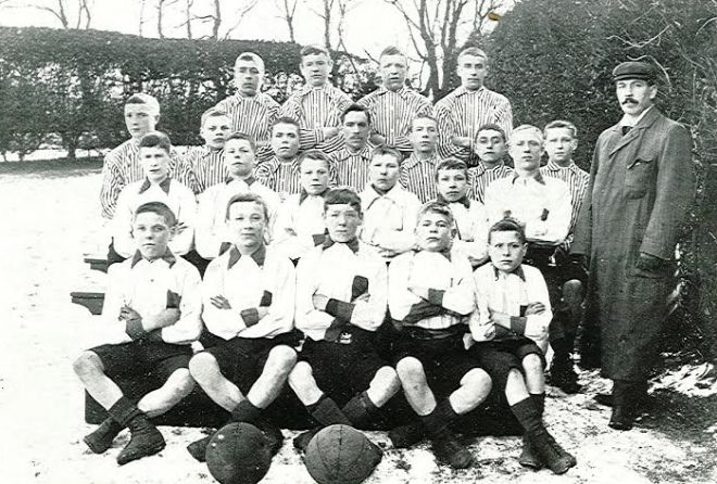 Black and white photo showing four lines of boys sitting with crossed arms and legs.  The first two lines wear white shirts, the back two lines wear striped shirts.  A man is standing by the side who is presumably the coach.