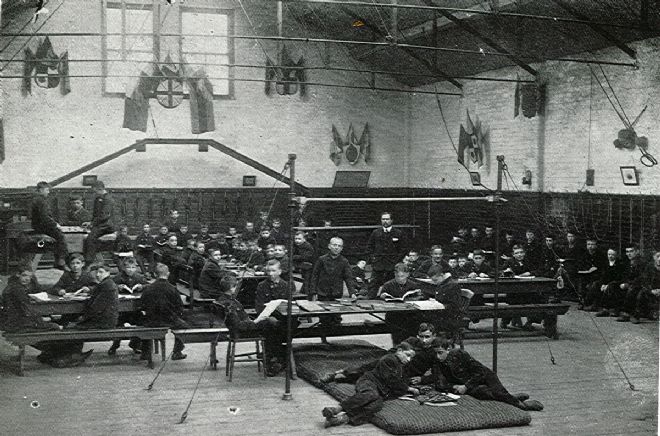 Black and white photo showing many boys in a gymnasium.  They are sitting at benches with tables with various gym equipment around.