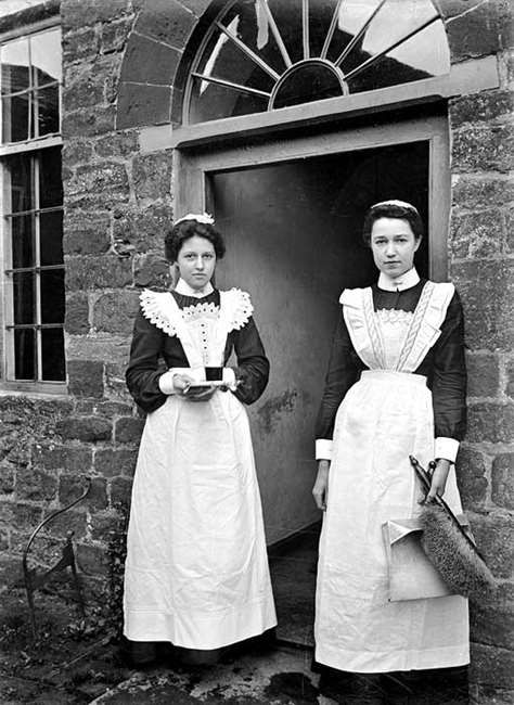 Two girls dressed in servant uniforms of black dresses with whtie pinafores over the top. One is holding a tray with a drink on it, the other is holding a brush.