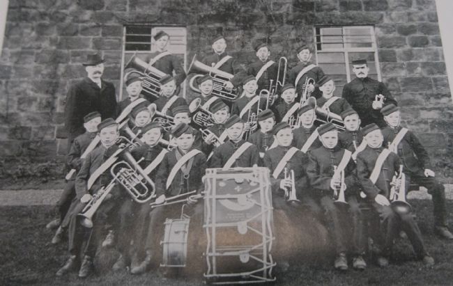 A group of boys with their brass instruments and two drums.  They are wearing uniforms with caps and a sash.