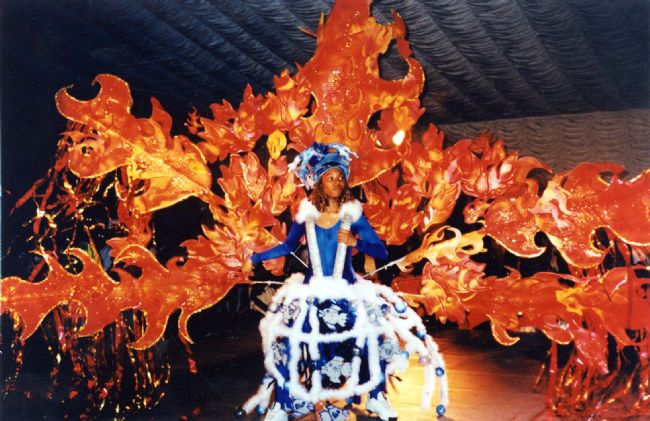 View of a young woman wearing a colourful and elaborate costume created for the Leeds West Indian Carnival. She is dressed in blue and is supporting a white 'cage' on her shoulders where fish appear to be swimming in and out. A huge canopy of flames in red, orange and gold fan out behind her.