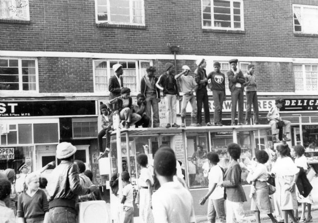 Crowds are gathered in Chapeltown Road on August Bank Holiday Monday, 25 August, 1980 to view the procession of the thirteenth West Indian Carnival to be held in Leeds since its beginnings in 1967. A group of youngsters have claimed a higher vantage point on top of a bus shelter.