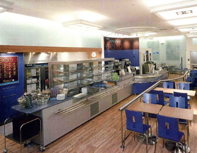 Colour photograph showing an empty cafe with a long self-service area, tables and blue plastic chairs
