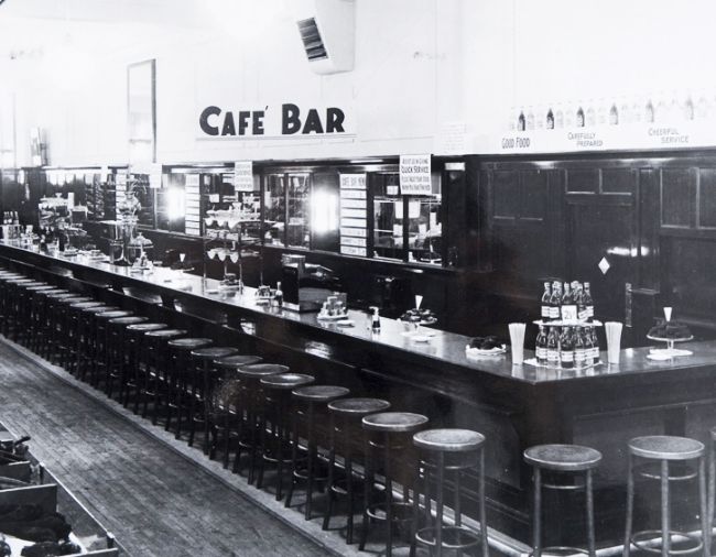 Black and white photo showing a long bar with bar stools