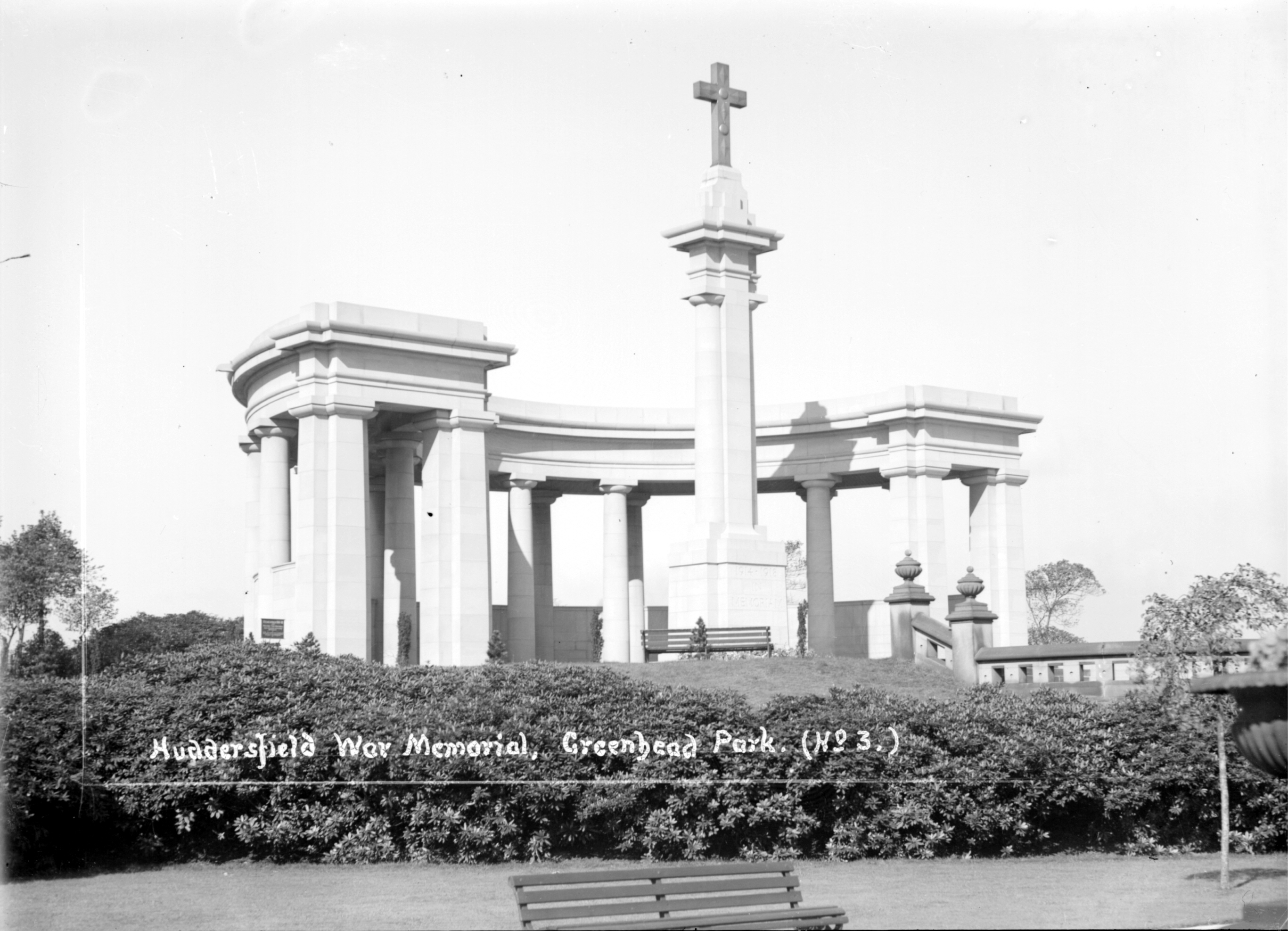 A black and white photograph of a white stone war memorial with tall cross, surrounded by trimmed bushes, benches, and flanked by trees under a clear sky. White text on the photograph reads 'Huddersfield War Memorial, Greenhead Park. (No 3.)'.