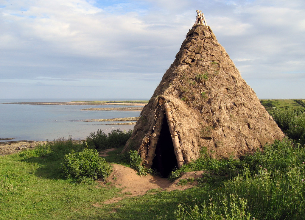 A reconstructed Mesolithic roundhouse with a conical shape made from wooden poles and thick thatch, standing on green grass beside a rocky coastline. The entrance is dark and narrow, with calm sea and a partly cloudy sky in the background.