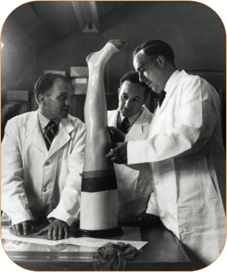 Three men in lab coats test a stocking for size on a plastic leg