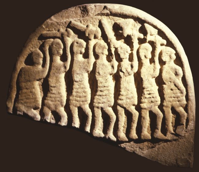 Stone carving picturing seven warriors brandishing axes above their heads.