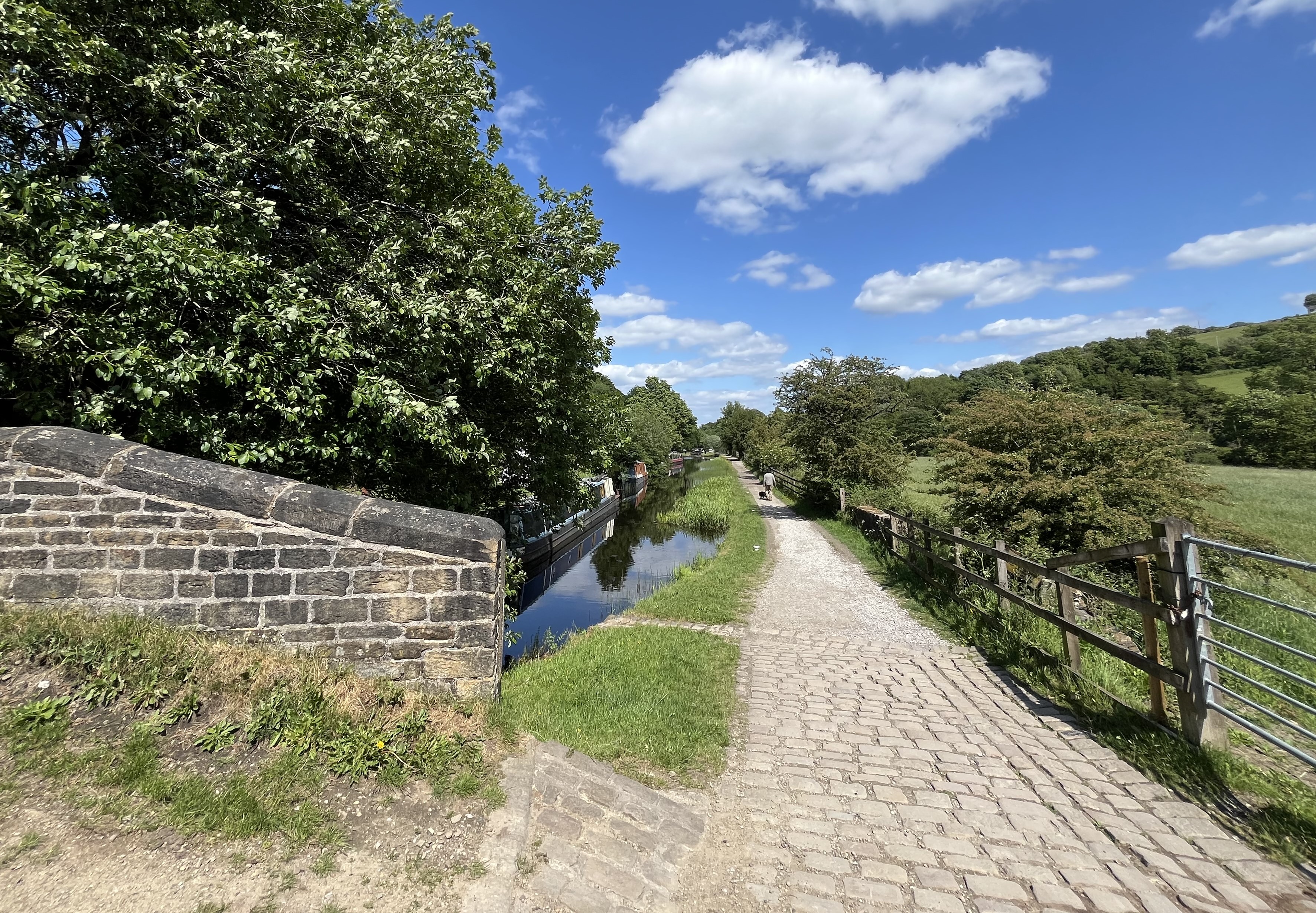 A scenic canal path bordered by lush green trees and a grassy field on a sunny day. There is a stone bridge in the foreground and cobbled path along the canal.